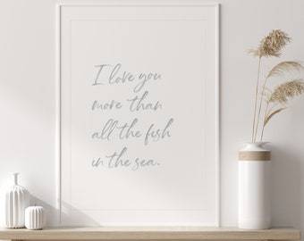 May include: A white framed print with the text "I love you more than all the fish in the sea." The print is hanging on a white wall above a wooden shelf with a white vase and a white ceramic object on it.
