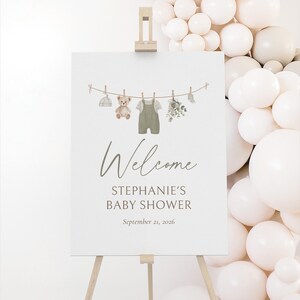 K&ouml;nnte beinhalten: Ein wei&szlig;es Schild auf einer Holzstaffelei mit der Aufschrift "Welcome STEPHANIE'S BABY SHOWER September 21, 2026". Das Schild zeigt eine Aquarellillustration von Babykleidung und einem Teddyb&auml;r, der an einer W&auml;scheleine h&auml;ngt.