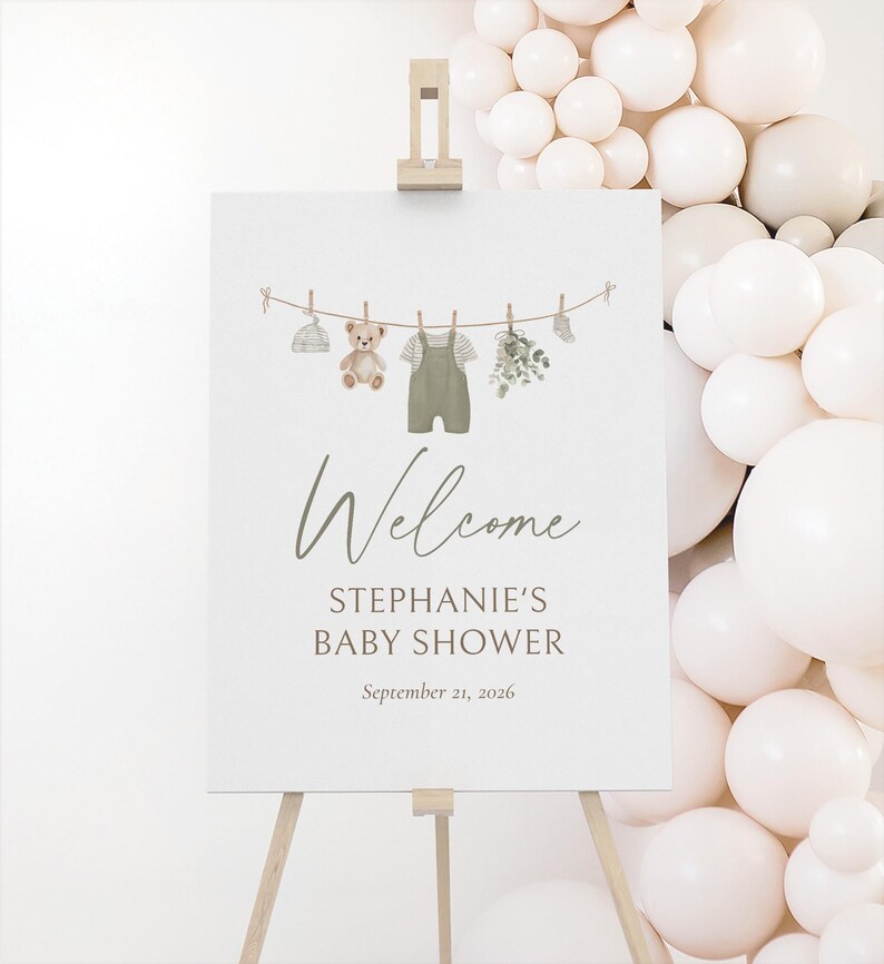 K&ouml;nnte beinhalten: Ein wei&szlig;es Schild auf einer Holzstaffelei mit der Aufschrift "Welcome STEPHANIE'S BABY SHOWER September 21, 2026". Das Schild zeigt eine Aquarellillustration von Babykleidung und einem Teddyb&auml;r, der an einer W&auml;scheleine h&auml;ngt.