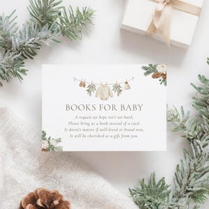 Puede incluir: Una tarjeta de invitación a un baby shower blanca con el texto "BOOKS FOR BABY" y una solicitud de libros en lugar de tarjetas. La tarjeta presenta ilustraciones de ropa y decoraciones para bebés, con ramas de pino y una caja de regalo.