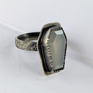 May include: A silver ring with a large, rectangular, gray gemstone. The gemstone is set in a textured bezel with a scalloped edge. The ring band features an ornate, engraved floral pattern.