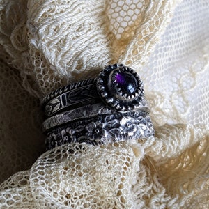 May include: A silver ring with a purple gemstone set in a bezel setting. The ring is stacked with three other silver bands, each with a different design. The ring is on a white lace background.