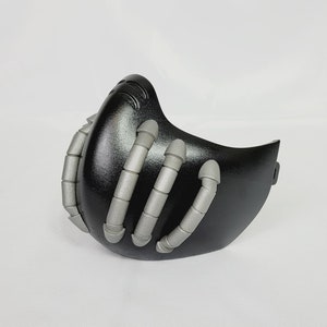 Puede incluir: Una máscara negra y plateada con un diseño único que presenta múltiples tubos plateados que sobresalen de la superficie. La máscara está hecha de un material liso y duro y tiene una forma ligeramente curvada.