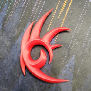 May include: A red plastic 3D printed Sonic the Hedgehog logo. The logo is a stylized design of a red flame with sharp points.