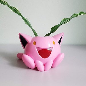 May include: A pink ceramic planter shaped like a cartoon character with large ears and a wide smile. The planter has a green plant growing out of the top.