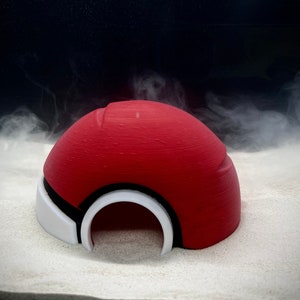 May include: A red and white Poke Ball shaped plastic hide for small pets, sitting on a white sandy surface.