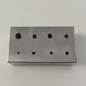 May include: A rectangular, silver-colored metal tool with eight circular indentations. Four larger indentations have a decorative, flower-like pattern. The other four are smaller and plain. The tool is likely used for crafting or jewelry making.