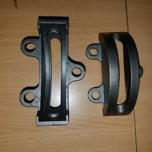 May include: Two black metal brackets with a curved shape and a rectangular opening. The brackets have multiple holes for mounting.
