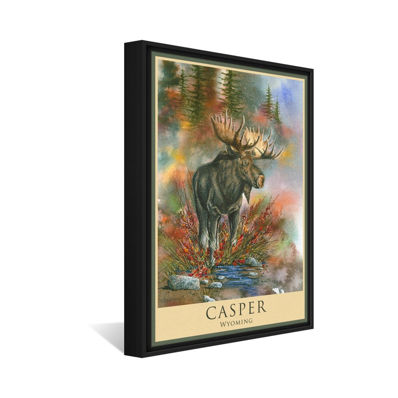 K&ouml;nnte beinhalten: Ein Aquarellbild eines Elchs, der in einem Bach steht, mit einem nebligen Wald im Hintergrund. Das Bild ist schwarz gerahmt und tr&auml;gt unten den Text "CASPER WYOMING".