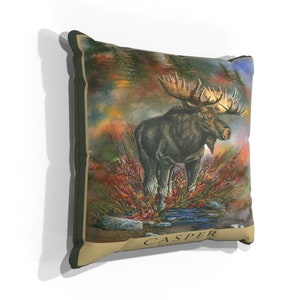 K&ouml;nnte beinhalten: Ein Dekokissen mit einer detaillierten Illustration eines Elchs, der in einer Waldlandschaft steht. Der Hintergrund ist ein Aquarellbild von B&auml;umen und Bl&auml;ttern in Gr&uuml;n-, Braun- und Oranget&ouml;nen. Der Elch ist braun mit gro&szlig;en Geweihen und steht in einem Wasserlauf. Das Kissen hat einen beigefarbenen Stoff mit den Worten "CASPER WYOMING" in Braun unten aufgedruckt.