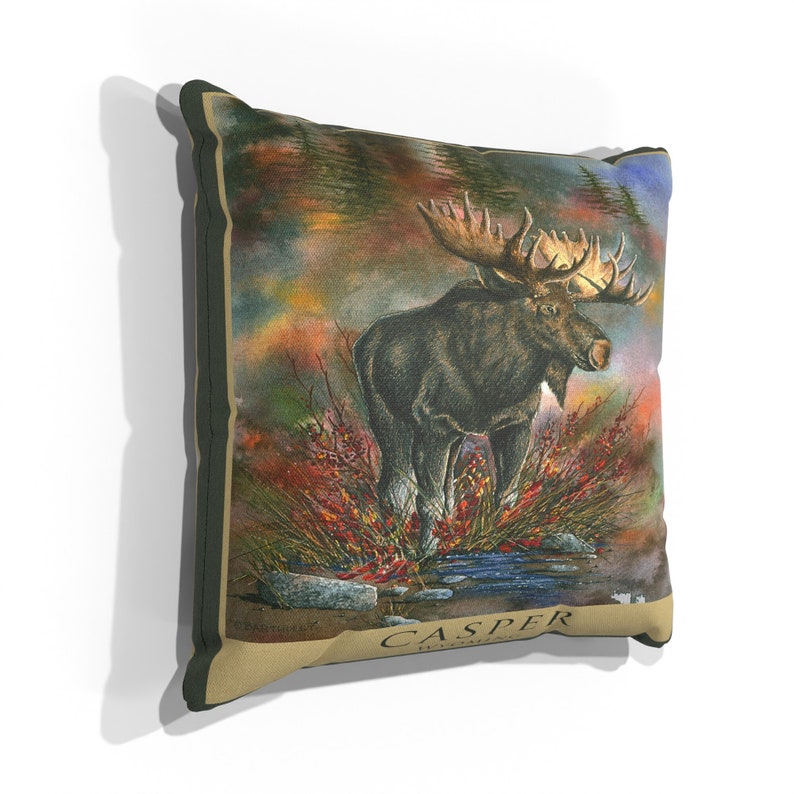 K&ouml;nnte beinhalten: Ein Dekokissen mit einer detaillierten Illustration eines Elchs, der in einer Waldlandschaft steht. Der Hintergrund ist ein Aquarellbild von B&auml;umen und Bl&auml;ttern in Gr&uuml;n-, Braun- und Oranget&ouml;nen. Der Elch ist braun mit gro&szlig;en Geweihen und steht in einem Wasserlauf. Das Kissen hat einen beigefarbenen Stoff mit den Worten "CASPER WYOMING" in Braun unten aufgedruckt.