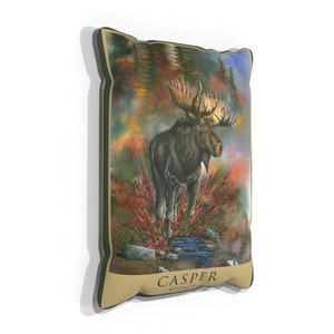 K&ouml;nnte beinhalten: Ein Dekokissen mit einem Aquarellbild eines Elchs, der in einem Wald mit einem Bach steht. Das Kissen hat einen braunen Hintergrund mit dem Text "CASPER WYOMING" unten.