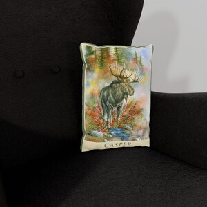 K&ouml;nnte beinhalten: Ein dekoratives Kissen mit einer Aquarell-Illustration eines Elchs, der in einem Bach steht. Der Hintergrund zeigt eine Waldszene mit B&auml;umen und Bl&auml;ttern. Das Kissen hat einen cremefarbenen Rand und das Wort "CASPER" ist in Schwarz unten aufgedruckt.