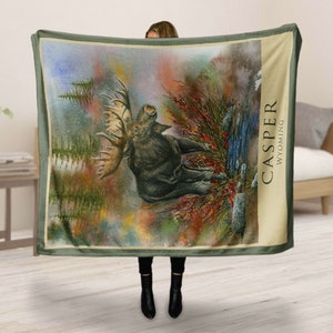 K&ouml;nnte beinhalten: Eine gr&uuml;ne Fleece-Decke mit einer Aquarell-Illustration eines Elchs in einer Waldlandschaft. Die Decke tr&auml;gt den Text "CASPER WYOMING" in der oberen rechten Ecke.