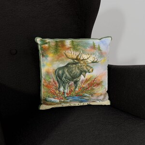 K&ouml;nnte beinhalten: Ein dekoratives Kissen mit einer Aquarell-Illustration eines Elchs, der auf einer Waldlichtung steht. Das Kissen hat einen braunen Hintergrund mit gr&uuml;nen B&auml;umen und roten Beeren. Der Elch ist braun mit gro&szlig;en Geweihen. Der Text "CASPER" ist auf der Unterseite des Kissens aufgedruckt.