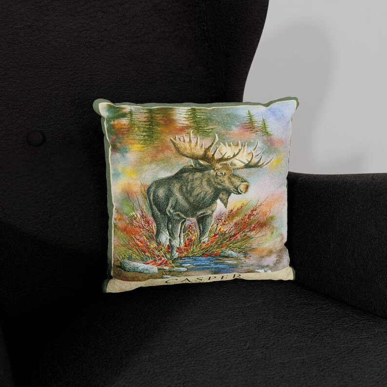 K&ouml;nnte beinhalten: Ein dekoratives Kissen mit einer Aquarell-Illustration eines Elchs, der auf einer Waldlichtung steht. Das Kissen hat einen braunen Hintergrund mit gr&uuml;nen B&auml;umen und roten Beeren. Der Elch ist braun mit gro&szlig;en Geweihen. Der Text "CASPER" ist auf der Unterseite des Kissens aufgedruckt.