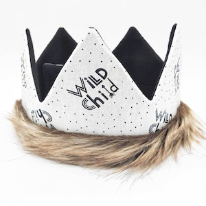 May include: A fabric crown with a white background, black polka dots, and the words "Wild Child" printed in black. The crown has black inner lining and a brown faux fur trim at the base. The crown is designed for children.
