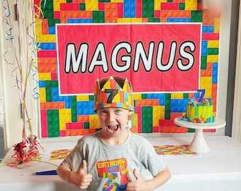May include: A birthday scene featuring a colorful backdrop with the name "Magnus" and a matching cake. A child wearing a crown with the number 7 and a gray t-shirt with the text "Birthday Boy" and "Magnus" gives a thumbs up.
