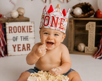 May include: A fabric birthday crown featuring a baseball pattern and the word ONE in red and white lettering. The crown has red fabric points at the top and is worn by a child sitting behind a white cake decorated with red baseball stitching.