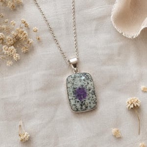 May include: A silver necklace with a rectangular pendant. The pendant features a speckled gray and white stone with a central purple design. The necklace is displayed on a cream-colored fabric, with dried flowers scattered around.