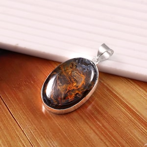 May include: Oval-shaped pendant with a polished, dark blue gemstone featuring swirling orange and brown patterns. The stone is set in a silver-colored metal frame with a matching bail for a necklace chain.