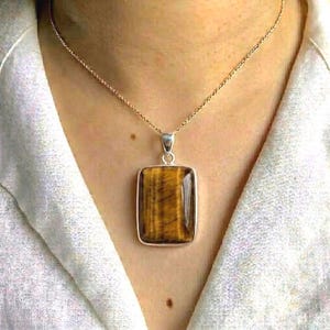 May include: A rectangular pendant necklace featuring a polished tiger's eye stone. The pendant is set in a silver-colored metal frame and hangs from a delicate chain. The tiger's eye stone displays a warm, golden-brown color with natural striations.