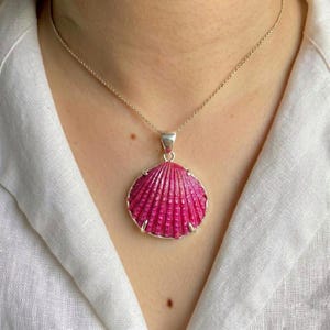 May include: A silver necklace featuring a vibrant pink seashell pendant. The pendant has a textured surface and is encased in a silver frame. The necklace is displayed against a white background.