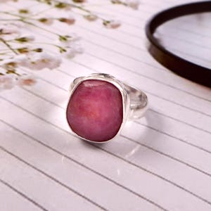 Peut inclure: Une bague en argent ornée d'une grosse pierre rose de forme irrégulière. La pierre est le point central, sertie dans un simple anneau en argent. La bague est présentée sur une surface blanche avec de fines lignes.