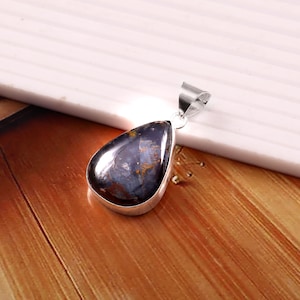 May include: A teardrop-shaped pendant with a polished, dark blue gemstone, featuring streaks of gold and brown. The stone is set in a silver-colored metal frame with a matching bail for a necklace chain. The pendant rests on a wooden surface.