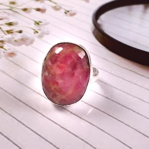May include: A silver ring featuring a large, oval-shaped, faceted pink gemstone. The gemstone has a mix of pink and red hues. The ring is set against a white striped background, with a blurred floral element in the upper left corner.