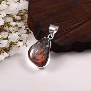 May include: A teardrop-shaped pendant with a silver bail. The pendant features a polished stone with a mix of dark blue, orange, and gold colors. The stone is set in a silver bezel. The pendant is likely a piece of jewelry.