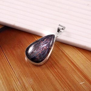 May include: A teardrop-shaped pendant with a polished, dark blue gemstone featuring red and brown veining. The stone is set in a silver-colored metal frame with a matching bail for a necklace chain.