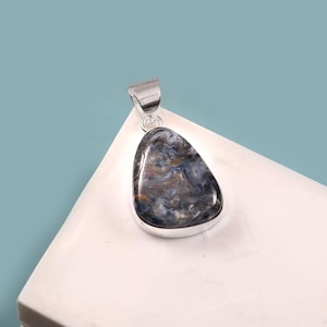 May include: A polished, teardrop-shaped pendant with a silver bail. The stone features a swirling pattern of blue, gray, and brown hues. The pendant is set in a silver bezel and rests on a white surface against a blue background.