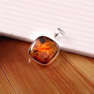 May include: A square pendant with a silver-colored frame. The pendant features a polished, amber-colored stone with orange and brown hues. The pendant is displayed on a wooden surface.