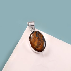 May include: Oval-shaped pendant with a polished tiger's eye stone, showcasing brown and dark blue swirls. The stone is set in a silver-colored metal frame with a matching bail for a necklace chain. The pendant rests on a white surface against a blue background.