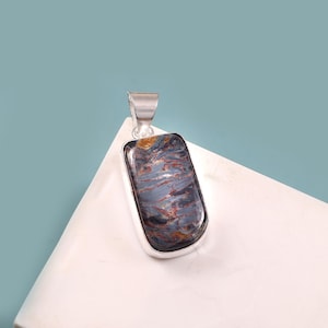 May include: A rectangular pendant with a silver bail. The pendant features a polished stone with a mix of blue, brown, and gray hues. The stone is set in a silver bezel. The pendant is displayed on a white surface against a blue background.