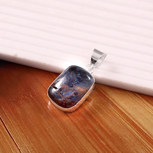 May include: A rectangular pendant with a polished, dark blue and brown stone set in a silver-colored metal frame. The pendant has a loop at the top for a chain. The stone's surface has a marbled appearance.