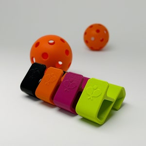 May include: A collection of colorful pickleball paddle clips in black, orange, magenta, and neon green, each featuring a paddle and ball design. Two orange pickleballs are also visible. The clips are designed to hold a paddle.
