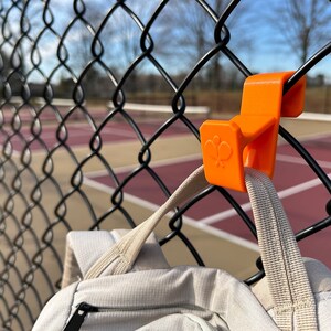 Puede incluir: Un gancho de pl&aacute;stico naranja est&aacute; sujeto a una valla de alambre negra, sosteniendo una mochila de color claro. El gancho tiene un logotipo estilizado. El fondo muestra una cancha de tenis.