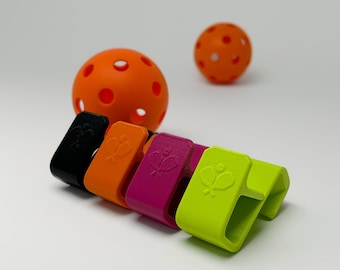 May include: A collection of colorful pickleball paddle clips in black, orange, magenta, and neon green, each featuring a paddle and ball design. Two orange pickleballs are also visible. The clips are designed to hold a paddle.