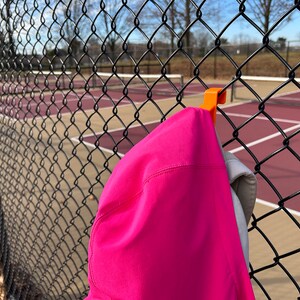 Puede incluir: Una chaqueta con capucha rosa brillante cuelga de un gancho naranja sujeto a una valla de alambre negra. El fondo muestra una cancha de tenis con l&iacute;neas rojas y blancas. La chaqueta es el foco principal de la imagen.
