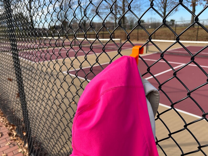 Puede incluir: Una chaqueta con capucha rosa brillante cuelga de un gancho naranja sujeto a una valla de alambre negra. El fondo muestra una cancha de tenis con l&iacute;neas rojas y blancas. La chaqueta es el foco principal de la imagen.
