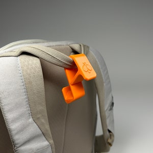 Puede incluir: Un clip naranja para mochila con un logotipo de mariposa est&aacute; sujeto a una correa de mochila gris. El clip es de pl&aacute;stico y tiene un dise&ntilde;o curvo. La mochila es de color gris claro con un patr&oacute;n texturizado.