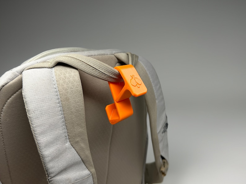 Puede incluir: Un clip naranja para mochila con un logotipo de mariposa est&aacute; sujeto a una correa de mochila gris. El clip es de pl&aacute;stico y tiene un dise&ntilde;o curvo. La mochila es de color gris claro con un patr&oacute;n texturizado.