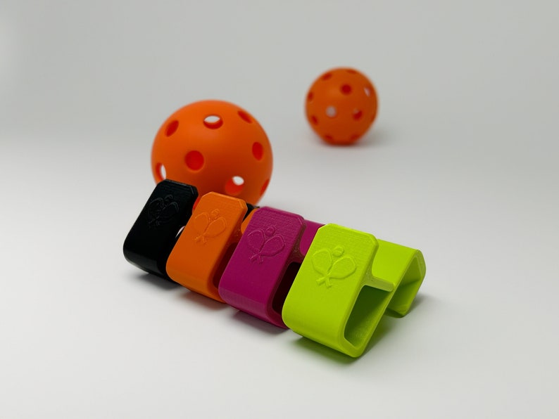 Puede incluir: Una colecci&oacute;n de clips coloridos para palas de pickleball en negro, naranja, magenta y verde ne&oacute;n, cada uno con un dise&ntilde;o de pala y pelota. Tambi&eacute;n se ven dos pelotas de pickleball naranjas. Los clips est&aacute;n dise&ntilde;ados para sujetar una pala.