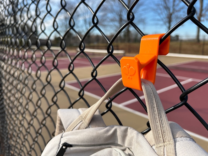 Puede incluir: Un gancho de pl&aacute;stico naranja est&aacute; sujeto a una valla de alambre negra, sosteniendo una mochila de color claro. El gancho tiene un logotipo estilizado. El fondo muestra una cancha de tenis.