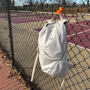 Puede incluir: Una mochila gris claro con el logotipo de North Face colgada de una valla de alambre negra. La mochila tiene un compartimento principal con cremallera y un bolsillo delantero. Se utiliza un gancho naranja para colgar la mochila.
