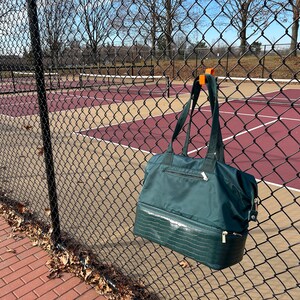 Puede incluir: Un bolso tote verde oscuro con una secci&oacute;n inferior texturizada cuelga de una valla de alambre. El bolso tiene cierre de cremallera y correas negras. Una cancha de tenis es visible en el fondo.