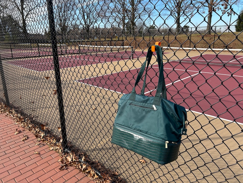 Puede incluir: Un bolso tote verde oscuro con una secci&oacute;n inferior texturizada cuelga de una valla de alambre. El bolso tiene cierre de cremallera y correas negras. Una cancha de tenis es visible en el fondo.