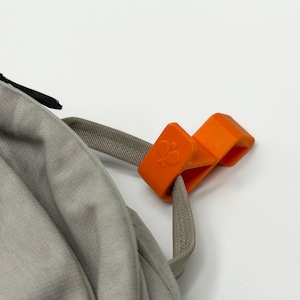 Puede incluir: Un clip naranja para mochila con un logotipo de mariposa est&aacute; sujeto a una correa gris y a una mochila gris. El clip est&aacute; hecho de un material pl&aacute;stico duro y tiene forma rectangular.